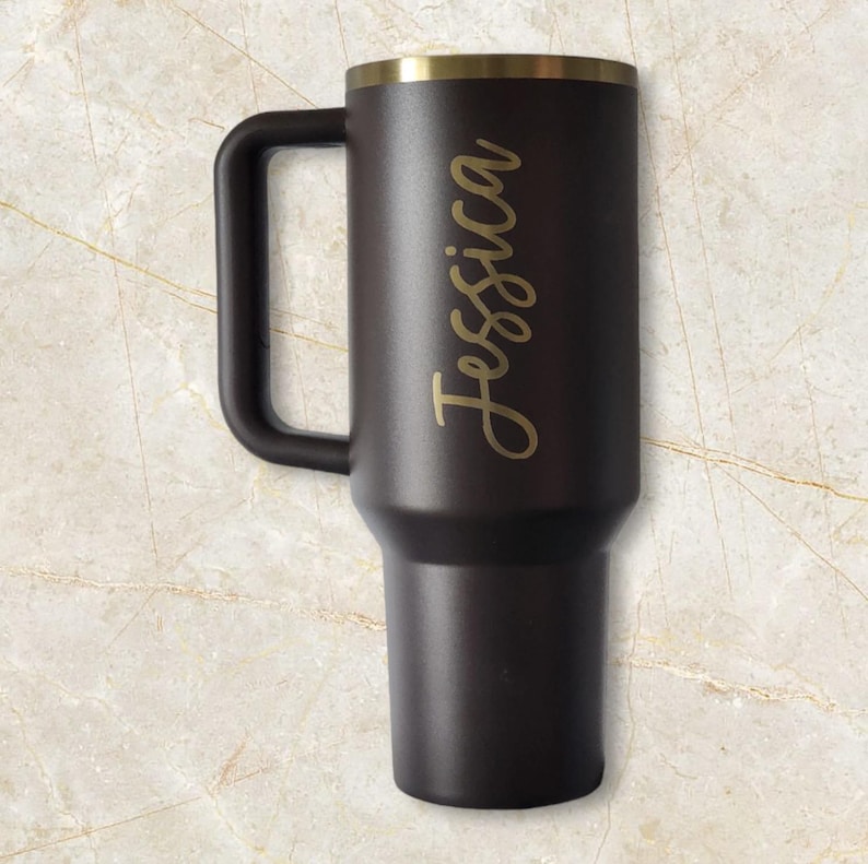 Puede incluir: Un termo de viaje negro con asa y borde dorado. El nombre "Jessica" est&aacute; escrito en letras doradas en el lateral. El termo est&aacute; dise&ntilde;ado para mantener las bebidas calientes o fr&iacute;as.