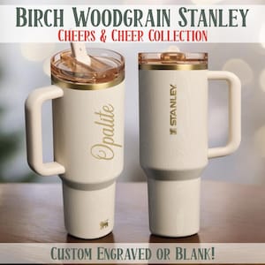 May include: Two cream-colored Stanley tumblers with gold accents and handles. One has "Opalite" in gold script, the other has the Stanley logo. The text "BIRCH WOODGRAIN STANLEY" and "CHEERS & CHEER COLLECTION" are at the top. The bottom reads "CUSTOM ENGRAVED OR BLANK!"