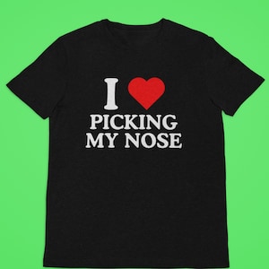 Puede incluir: Camiseta negra con la frase "I ❤️ PICKING MY NOSE" en texto blanco y un corazón rojo. La camiseta se muestra sobre un fondo verde.