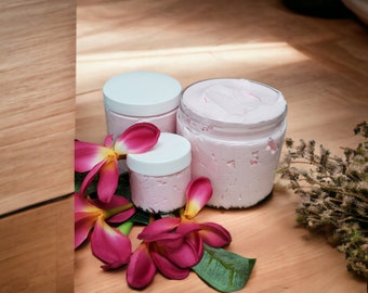 May include: Three jars of pink body butter with white lids. The jars are arranged in a pyramid shape on a wooden surface. There are pink flowers and dried lavender sprigs around the jars.