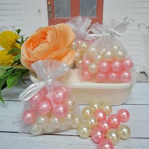 May include: Three small white organza bags filled with pink and white pearl beads. The bags are tied with white ribbon and are sitting on a white surface.