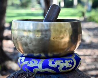 May include: A large, silver singing bowl with a wooden striker resting on the rim. The bowl is sitting on a blue and gold patterned cushion.