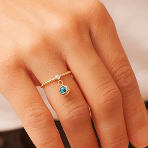 May include: A gold ring with a small blue evil eye charm dangling from it. The ring is set with a small clear stone.