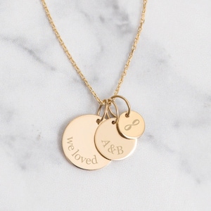 May include: Gold-colored pendant necklace featuring three circular charms. One charm is engraved with "we loved", another with "A&B", and the third with an infinity symbol. The necklace is displayed on a marble surface and worn by a person.