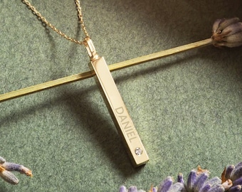 May include: A gold bar necklace with the name "DANIEL" engraved on it. The necklace is on a green background with lavender flowers.