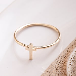 May include: A simple gold ring with a small cross pendant. The ring is on a white surface.