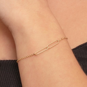 May include: A gold-colored jewelry set featuring a necklace and bracelet with a paperclip design. The bracelet is on the wrist, and the necklace is around the neck. A gold ring is on the finger. The jewelry is worn over a black tank top.
