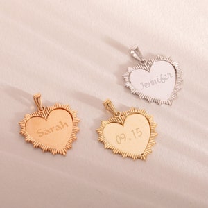 May include: Three gold heart-shaped pendants with different engravings. The pendants are on a light pink background. The engravings are "Sarah", "Jennifer", and "09.15".