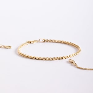 May include: A gold-toned box chain bracelet featuring a series of interconnected square links. The bracelet is circular in shape and includes a small metal clasp mechanism at one end, with additional chain segments extending from the main piece.