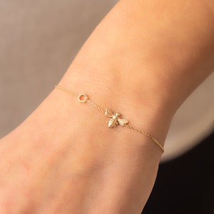 Könnte beinhalten: Zartes goldfarbenes Armband mit einem Bienen-Anhänger und einem sechseckigen Akzent. Das Armband wird am Handgelenk getragen und zeigt seine feinen Details und sein elegantes Design. Der Bienen-Anhänger verleiht dem Schmuck einen Hauch von Natur.