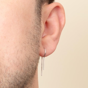 May include: A close-up of a person's ear with a silver earring. The earring is a thin, straight wire with a small loop at the top. The wire is bent at the bottom to create a small hook.