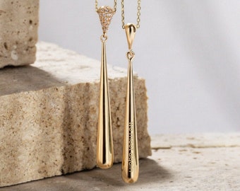 May include: Two gold-colored pendant necklaces. One features a teardrop-shaped pendant with the text "WE MEET AGAIN" engraved on it. The other has a triangular top encrusted with small, clear stones. Both hang from delicate gold chains.
