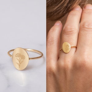 May include: A gold ring with a delicate oval-shaped design featuring an engraved floral motif. The ring is shown on a model's finger.