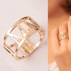 Pode incluir: Um anel dourado com uma banda larga que apresenta um design de numerais romanos. O anel &eacute; aberto e tem um design recortado.