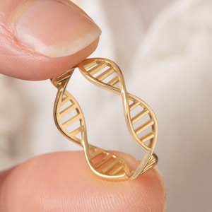 May include: A gold-coloured ring shaped like a DNA double helix. The ring is held between two fingers against a soft, white background. The ring's design features a repeating pattern of connected lines.