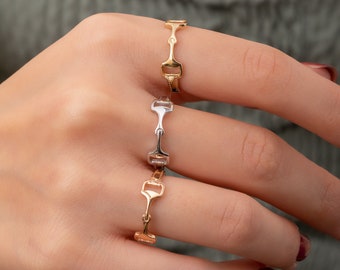 Puede incluir: Tres anillos usados en los dedos, con un diseño de bocado de caballo. El anillo superior es de oro amarillo, el del medio es de tono plateado y el inferior es de oro rosa. Cada anillo tiene una banda delgada y un motivo central de metal.