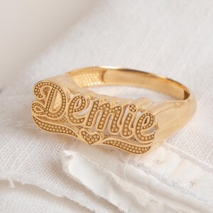 Può includere: Un anello dorato con il nome "Demie" in un carattere corsivo ornato. L'anello presenta un design a cuore sotto il nome ed è posto su uno sfondo di tessuto bianco. Un gioiello personalizzato.