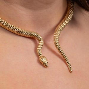 Puede incluir: Un collar de serpiente dorado con un patrón texturizado en forma de escamas. La cabeza de la serpiente está en la parte delantera, con su cuerpo curvándose alrededor del cuello. Este collar es una pieza llamativa.