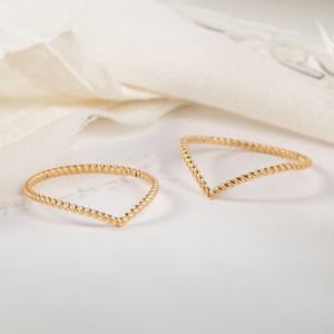 Könnte beinhalten: Zwei goldfarbene, V-förmige Ringe mit einem gedrehten Seildesign. Die Ringe sind vor einem weißen Hintergrund mit einem weichen, neutral getönten Stoff im Hintergrund platziert. Die Ringe sind zart und elegant.