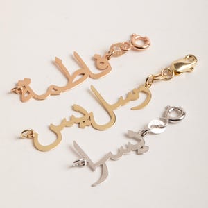 Può includere: Tre ciondoli con nome in metallo dorato, oro rosa e argento. Ogni ciondolo presenta un nome in scrittura araba con un moschettone per un facile fissaggio a un braccialetto o una collana. I ciondoli sono esposti su sfondo bianco.