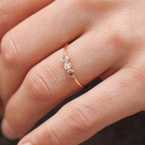 Può includere: Un sottile anello in oro con tre piccole pietre preziose rotonde incastonate a lunetta nei colori viola, trasparente e rosa. Sotto l'anello, una tabella etichettata BRITHSTONE mostra dodici pietre circolari colorate per ogni mese dell'anno.