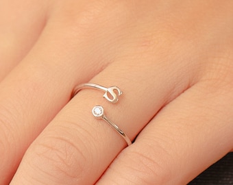Può includere: Un anello d'argento con un piccolo diamante e una lettera 'S' sulla fascia. L'anello è indossato su un dito con smalto verde.
