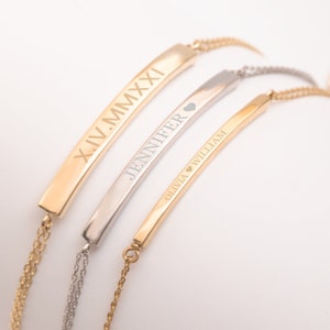 Könnte beinhalten: Drei goldene und silberne Bar-Armbänder mit eingraviertem Text. Das obere Armband ist golden und trägt die Aufschrift "XIV.MMXXI". Das mittlere Armband ist silberfarben und trägt die Aufschrift "JENNIFER". Das untere Armband ist golden und trägt die Aufschrift "OLIVIA ♥ WILLIAM".