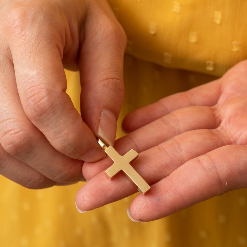 May include: A person's hand holding a gold cross pendant. The cross is simple and has a smooth, polished finish.