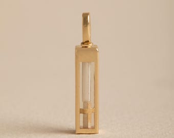 May include: A rectangular gold pendant featuring a vertical clear glass chamber. Inside the chamber, a white cylindrical element sits above a small, textured gold cross. The pendant has a polished gold frame and a matching gold bail at the top.
