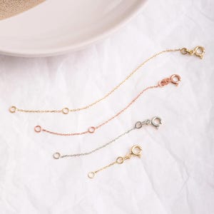 May include: Three gold chain extenders in different lengths and colours: gold, rose gold, and silver. The extenders are on a white surface.
