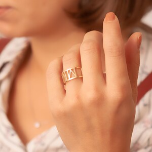 A close-up of a hand wearing a yellow gold ring with a distinctive cut-out Roman numeral design, highlighting its elegant fit and craftsmanship. The blurred background emphasizes the ring's intricate details, showcasing its suitability as a fashionable jewelry.