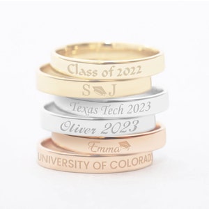 May include: Stack of five gold, silver, and rose gold rings with engraved graduation dates and text. The top ring says "Class of 2022" and "S.J." with a graduation cap symbol. The next ring says "Texas Tech 2023". The third ring says "Oliver 2023". The fourth ring says "Emma" with a graduation cap symbol. The bottom ring says "University of Colorado".
