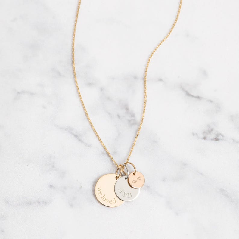 May include: A gold-colored necklace with three pendant charms. The largest charm is a gold disc with the engraved words "we loved". The other charms are a silver disc with "A&B" and a small rose gold charm with an infinity symbol.
