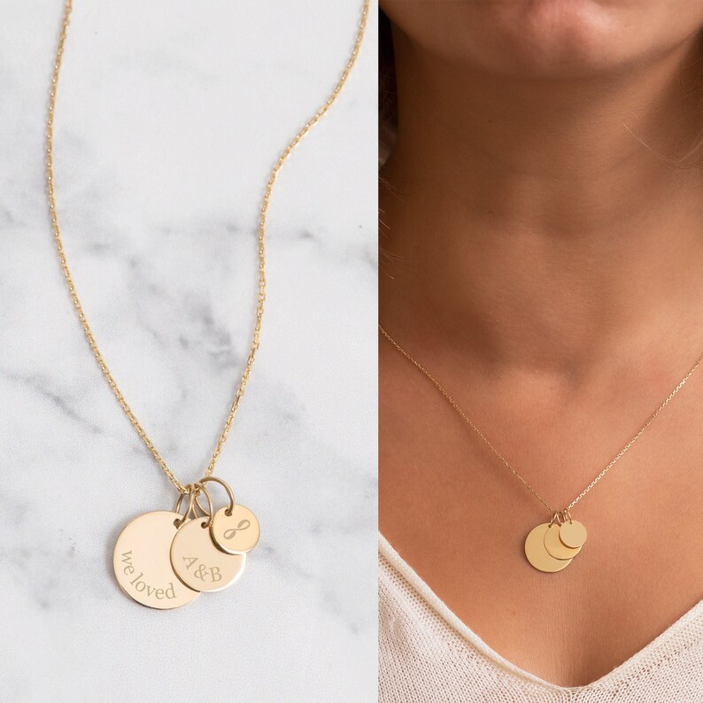 May include: Gold-colored pendant necklace featuring three circular charms. One charm is engraved with "we loved", another with "A&B", and the third with an infinity symbol. The necklace is displayed on a marble surface and worn by a person.