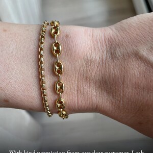 May include: Two gold-toned bracelets worn together. One bracelet features a delicate, rounded box chain design, while the other consists of larger, oval-shaped links with a central bar, creating a layered look on the wrist.