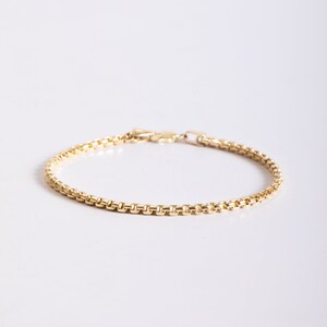 May include: A gold-toned box chain bracelet featuring a series of interconnected square links that form a smooth, continuous rounded profile. The bracelet includes a small, secure lobster claw clasp mechanism at the end for fastening the piece.