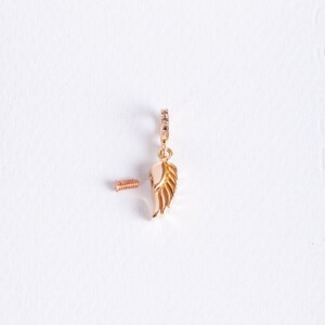 K&ouml;nnte beinhalten: Ein goldfarbener Anh&auml;nger mit einem detaillierten Fl&uuml;geldesign und einer kleinen Schraube zur Befestigung. Der Anh&auml;nger hat einen kleinen Kreis aus klaren Steinen um die Oberseite.