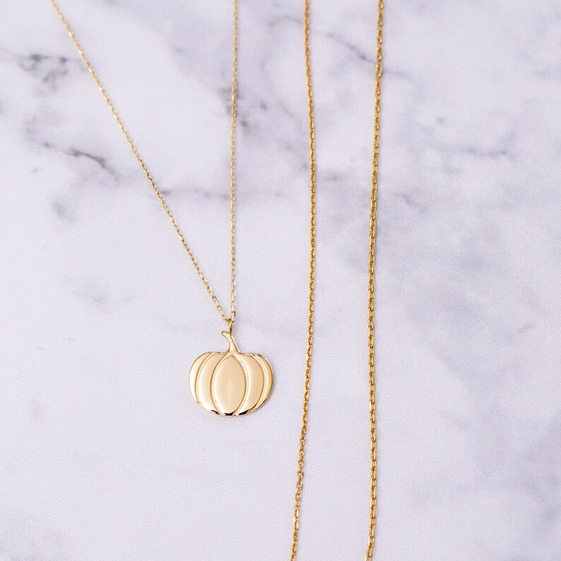 Puede incluir: Un collar de cadena de oro con un colgante de calabaza. El collar est&aacute; sobre una superficie de m&aacute;rmol blanco.
