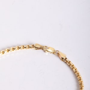 May include: A gold-toned box chain necklace featuring a polished finish and a secure lobster claw clasp. The chain consists of interconnected square links that create a smooth, uniform appearance. The clasp mechanism is clearly visible and functional.