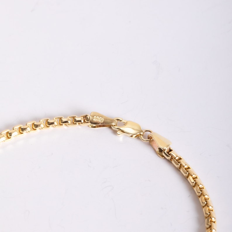 May include: A gold-toned box chain necklace featuring a polished finish and a secure lobster claw clasp. The chain consists of interconnected square links that create a smooth, uniform appearance. The clasp mechanism is clearly visible and functional.