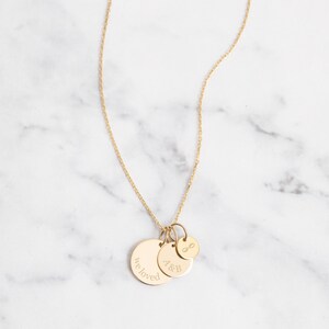 May include: A gold-colored necklace with three pendant charms. The largest charm has the words "we loved" engraved on it. The middle charm has the letters "A&B" engraved. The smallest charm has an infinity symbol. The necklace is on a white marble surface.