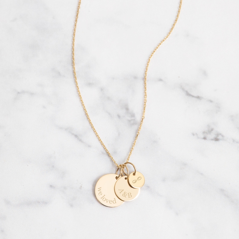 May include: A gold-colored necklace with three pendant charms. The largest charm has the words "we loved" engraved on it. The middle charm has the letters "A&B" engraved. The smallest charm has an infinity symbol. The necklace is on a white marble surface.