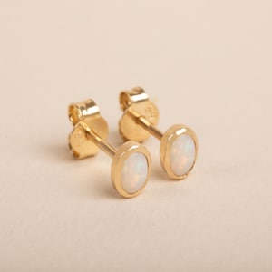 May include: A pair of gold stud earrings featuring oval-shaped white opals set in polished gold bezels. The earrings include standard gold butterfly-style friction backs, showcasing a simple design with iridescent gemstone centres.
