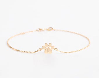May include: A gold-toned chain bracelet featuring a central paw print charm. The charm is engraved with the capital letter K in the center. The bracelet is constructed with a delicate link chain and includes a small metal clasp for closure.