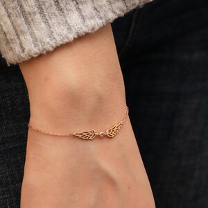Puede incluir: Una delicada pulsera dorada con un par de alas estilizadas con intrincados detalles de plumas. Una peque&ntilde;a gema rosa est&aacute; engastada entre las alas. La pulsera se lleva en una mu&ntilde;eca, con un su&eacute;ter gris y vaqueros oscuros en el fondo.
