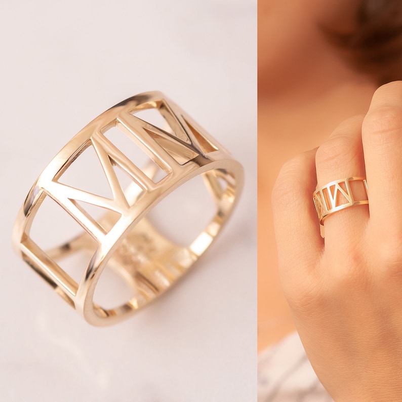 Pode incluir: Um anel dourado com uma banda larga que apresenta um design de numerais romanos. O anel &eacute; aberto e tem um design recortado.