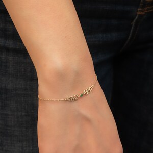 Puede incluir: Pulsera dorada con dise&ntilde;o de alas en cada lado, unidas por una fina cadena. Una peque&ntilde;a gema verde se encuentra entre las alas. La pulsera se lleva en la mu&ntilde;eca.