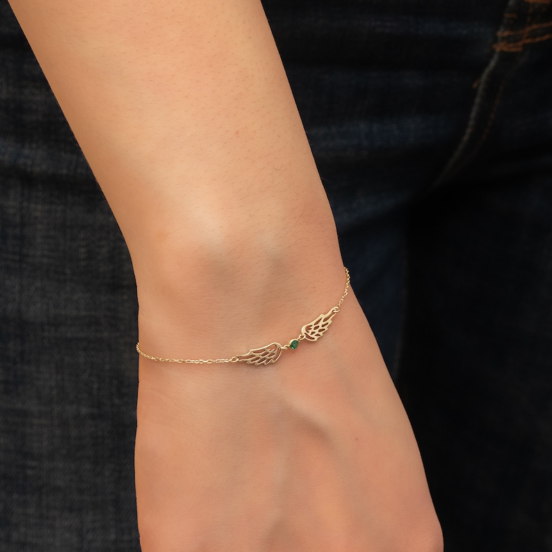 Puede incluir: Pulsera dorada con dise&ntilde;o de alas en cada lado, unidas por una fina cadena. Una peque&ntilde;a gema verde se encuentra entre las alas. La pulsera se lleva en la mu&ntilde;eca.