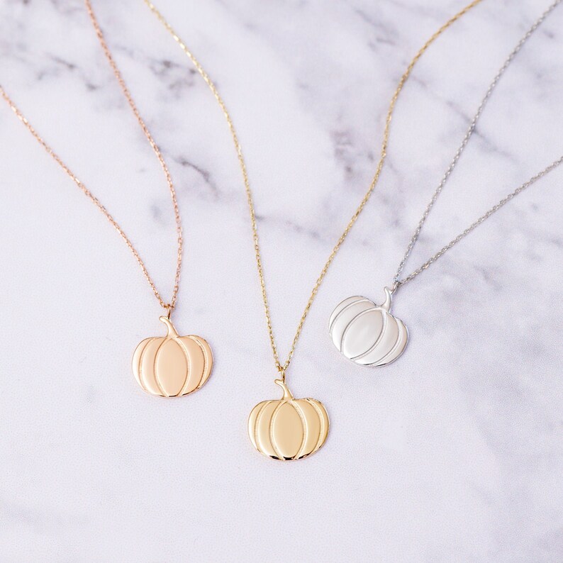 Puede incluir: Tres collares de oro con colgantes de calabaza. Los collares est&aacute;n sobre una superficie de m&aacute;rmol blanco. Las calabazas son de diferentes tonos de oro.