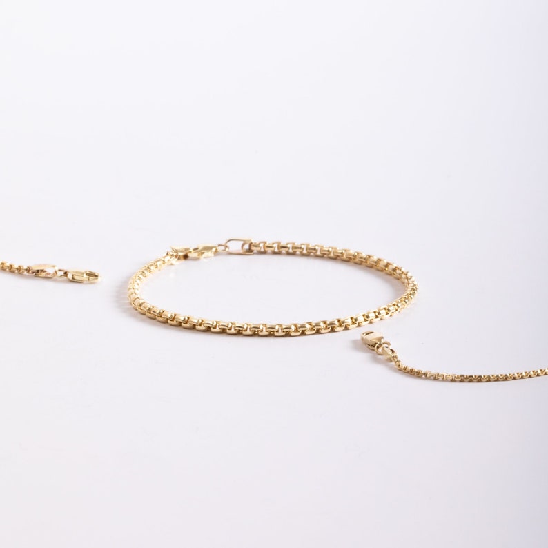May include: A gold-toned box chain bracelet featuring a series of interconnected square links. The bracelet is circular in shape and includes a small metal clasp mechanism at one end, with additional chain segments extending from the main piece.